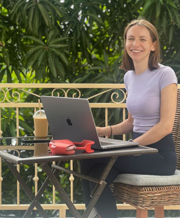 Eine Kollegin sitzt lächelnd mit einem Laptop an einem kleinen Tisch auf einer Terrasse, im Hintergrund dichte grüne Pflanzen. Neben dem Laptop steht ein Iced Coffee. Die Szene zeigt entspanntes Remote-Arbeiten im Freien.